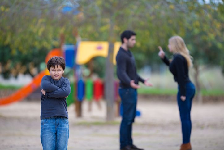 A young boy sits on a playground bench holding his head while his parents argue in the background, illustrating the stress of child custody conflict.